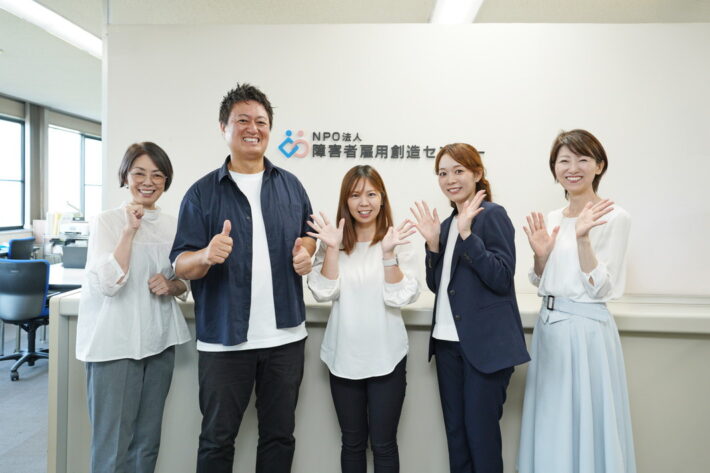 事業所の入口で温かく出迎える5人のスタッフ。誰もが自分らしく輝ける場所として、相談しやすい雰囲気と笑顔を絶やさないチームワークの象徴