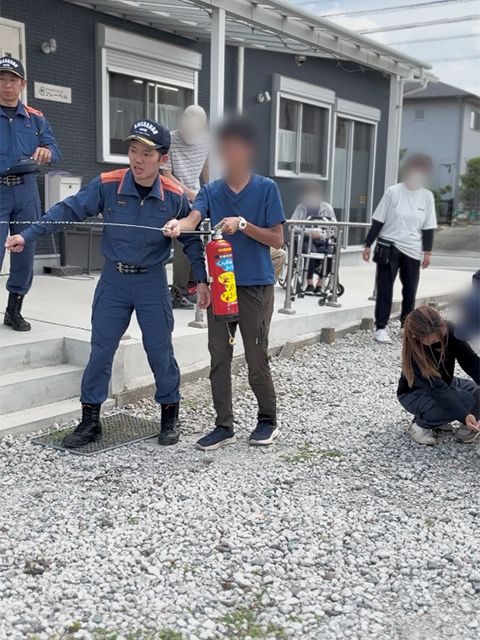 フレーベル施設内での消防訓練にて、消防署員から指導を受けながら、水消火器のピンを抜き、ホースを構えて初期消火の基本を学ぶ練習。
