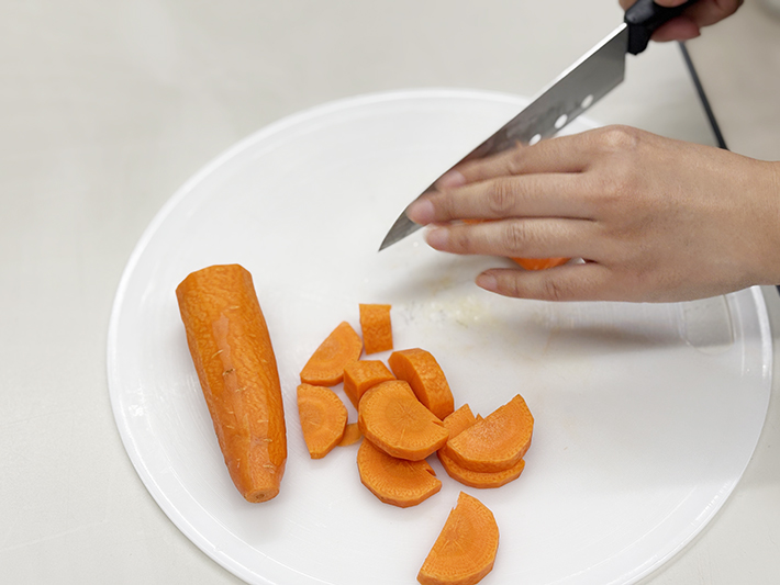 訓練生が慎重に包丁を使い、カレーの具材となるニンジンを均等な厚さの半月切りにカットしている手元のアップ。