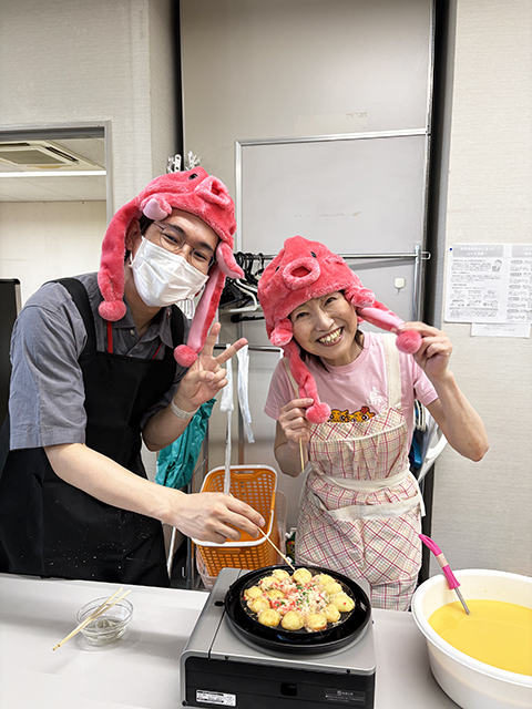 たこ焼き器を囲み、実演を行う調理スタッフ。美味しそうな香りが漂う中、焼き方のコツを訓練生に伝える重要なシーン