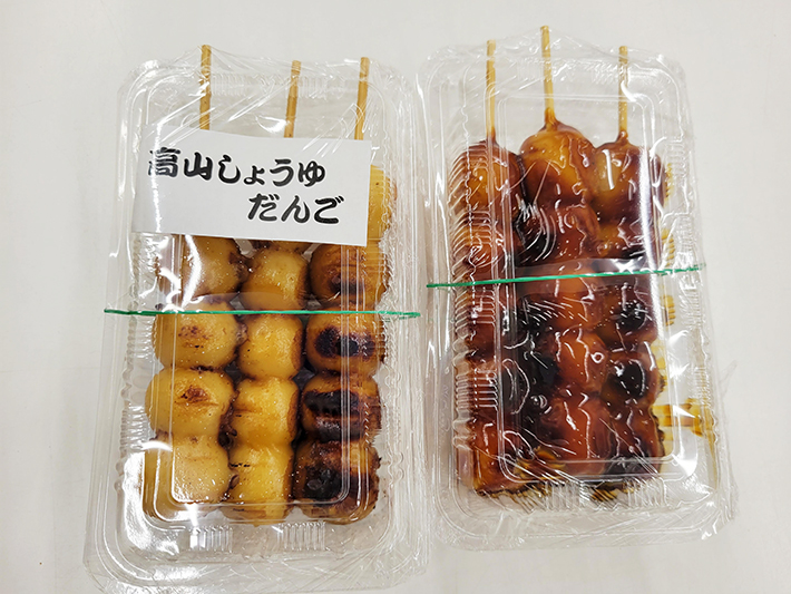 「花より団子」を象徴する、香ばしそうな2種類の団子。左側には「高山しょうゆだんご」のラベルが添えられ、梅雨の時期に室内で楽しむ美味しいひとときを表現。