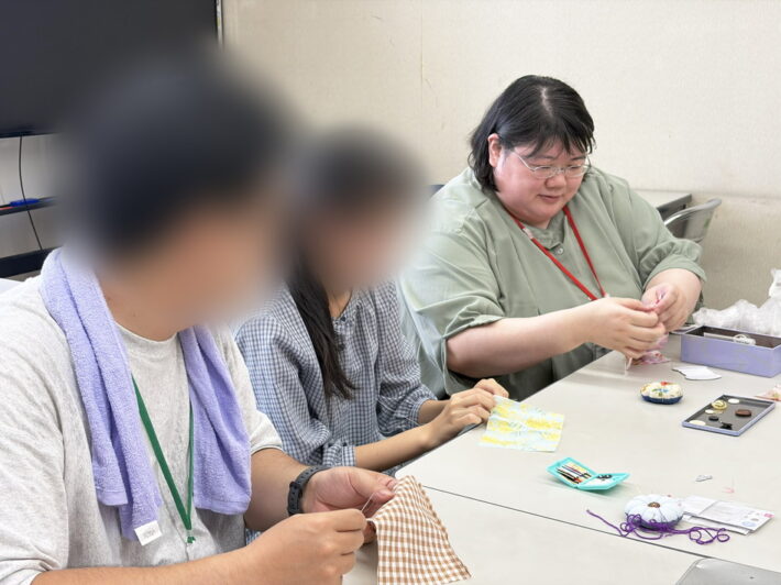 スタッフが実演する「玉結び」や「並縫い」の指使いを間近で見学し、正しい針の運び方や布の持ち方を確認しながら習得を目指す訓練生。
