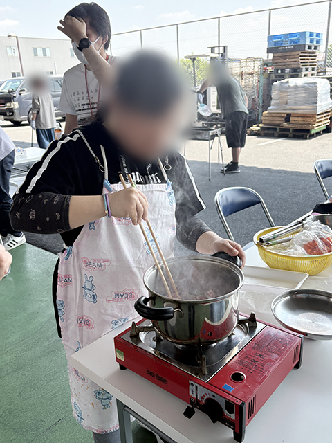 大鍋を火にかけ、炒めた肉や野菜を煮込んでいる野外調理のメインシーン。