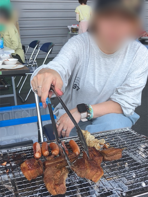 渡邉理事特製の柔らかそうな豚の角煮をトングで取り分け、横でウインナーも焼いているBBQの風景。