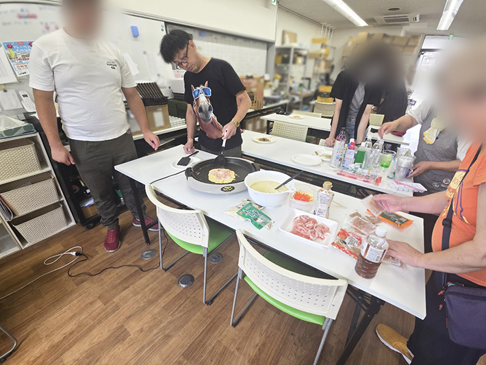 ホットプレートでお好み焼きの準備を進めるオリーブのスタッフ。豚肉やキャベツなどの具材を乗せ、香ばしい匂いが立ち込める中、訓練生の皆さんがバトンタッチしてひっくり返すのを待っている場面。