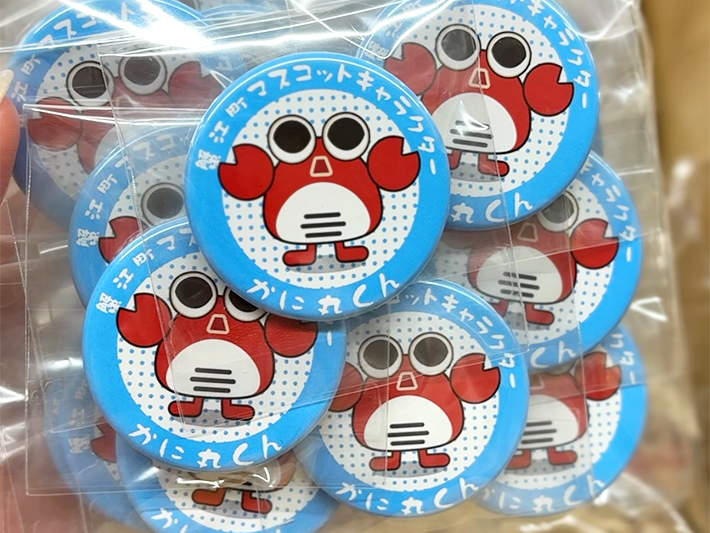 蟹江町観光協会様よりご依頼いただいた、蟹江町マスコットキャラクター「かに丸くん」の缶バッジ。ブロアーでほこりを除去し、専用のメーカーで組み立て・検品を行った後、一つずつ丁寧にOPP袋へ封入・封緘(ふうかん)された完成品の様子。