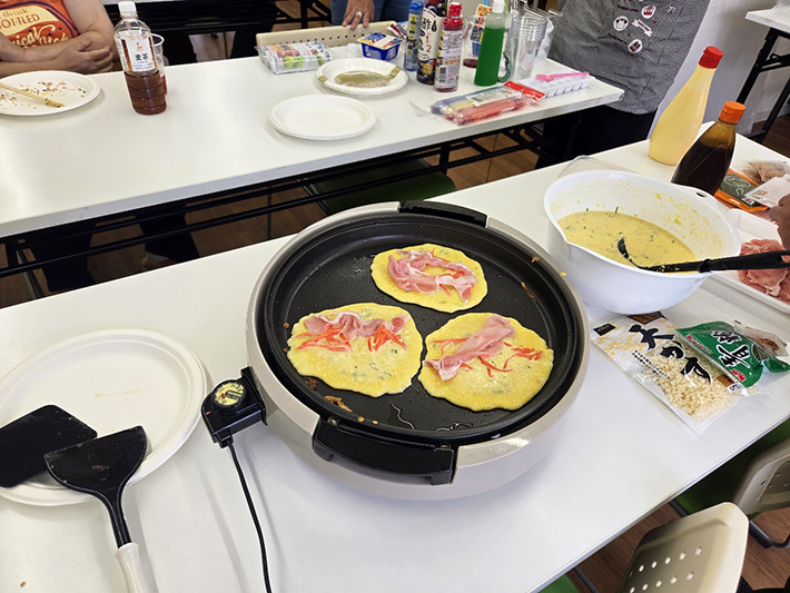 ホットプレート上の生地に、色鮮やかな紅しょうがや新鮮な豚肉がトッピングされた様子。具だくさんで栄養満点なお好み焼きが、美味しそうな音を立てて焼き上がっていく調理のプロセス。