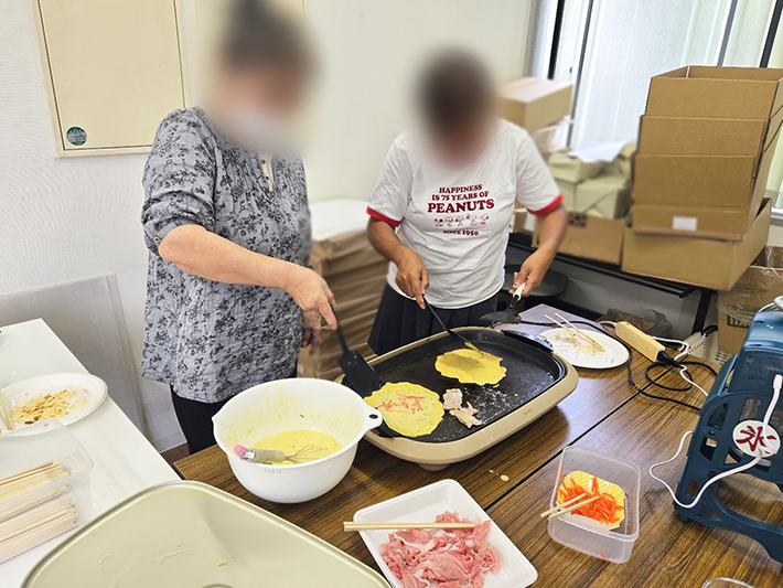 「もう焼けたかな？」とお好み焼きの裏側を少し持ち上げ、焼き色を慎重に確認する訓練生。丁寧な作業を心がけ、最高の一枚を焼き上げようとするこだわりが伝わる瞬間。