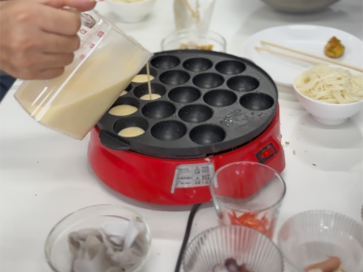 グループごとに分かれた訓練生たちが、協力してたこ焼きの生地をプレートの穴へ丁寧に流し入れている場面。役割分担をしっかり行い、チームワークを発揮しながら作業を進める訓練の一環。
