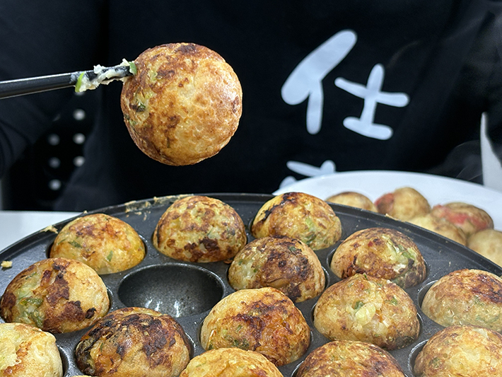 熱々のたこ焼きを一粒つまみ上げる様子。自分たちで協力して作り上げた「格別のおいしさ」を実感し、達成感を分かち合うひととき。