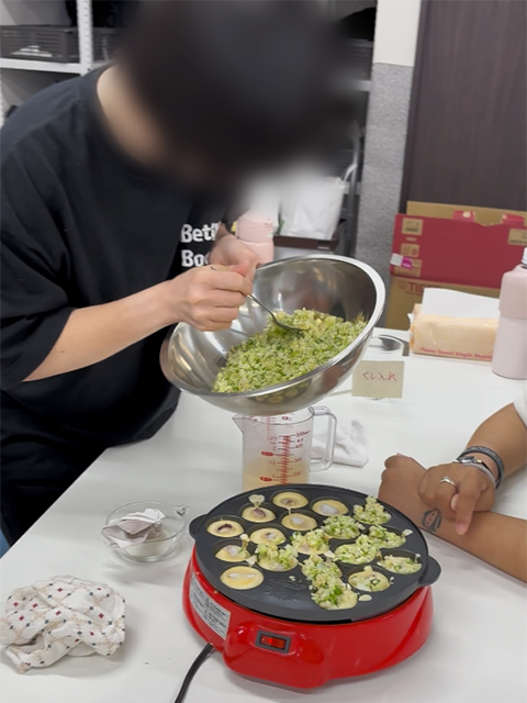 たこ焼き器いっぱいに広げた具材を整え、たこ焼きの形を作る準備をする様子。利用者同士で自然と会話が生まれ、笑顔があふれるフレーベル恒例のレクリエーション風景。
