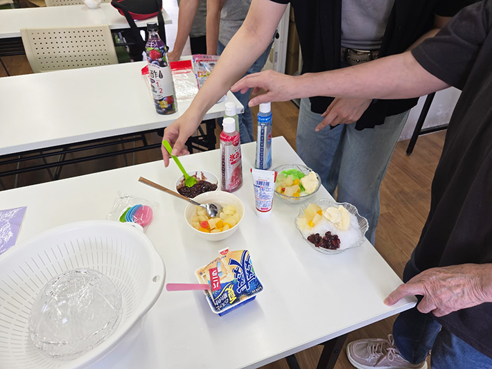 かき氷の彩りを添えるあんこ、フルーツポンチ、バニラアイスなどの豪華なトッピングのラインナップ。自由に選べる楽しさが、夏のレクリエーションをさらに盛り上げている様子。