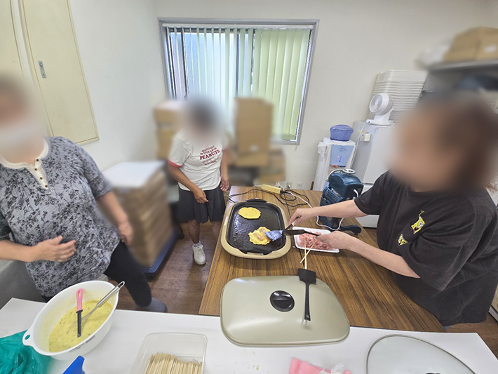 へらを使ってお好み焼きの形を整え、軽く押し付けて焼き固める訓練生。調理を通じて火加減の確認や手先の動作を学び、自分たちで作り上げる達成感を味わっている風景。