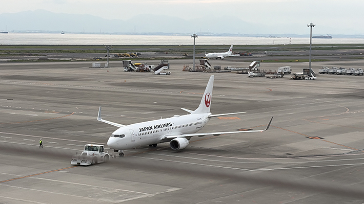 滑走路の手前で離陸許可を待つ日本航空（JAL）の旅客機。尾翼の鶴丸ロゴが誇らしく、エンジンの音がスカイデッキまで響いてきそうな臨場感のある一枚。