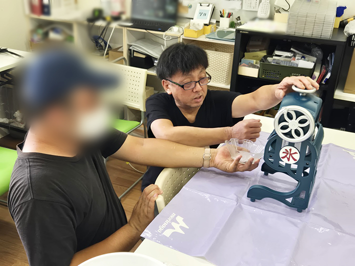かき氷機を操作し、出来立てのふわふわな氷を訓練生のお椀にふるまうスタッフ。利用者とスタッフの笑顔の対話が生まれる、オリーブらしい温かなコミュニケーションの時間。