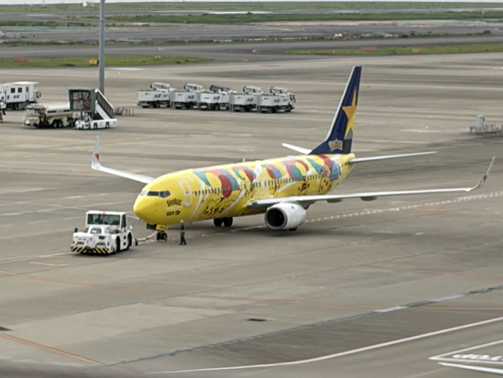 駐機場でトーイングカー（牽引車）に引かれ、ゆっくりと誘導路へ移動するスカイマークの「ピカチュウジェット」。鮮やかな黄色の機体が、空港の機能美の中で一際目を引く様子。