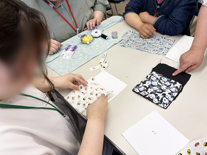 テーブルに並べられた様々な色や柄の布地から、お気に入りのデザインを真剣に選ぶ訓練生。「この可愛い絵柄が表側に綺麗に出るようにしたい」と、完成後のポーチを具体的にイメージしながら楽しく思索する様子。