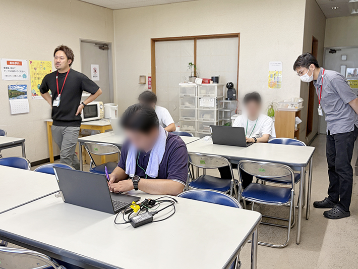 障碍者ITカレッジ愛西で毎月開催されている「生成AI活用講座」にて、パソコンに向かって真剣にプロンプトを組み立てる訓練生と、優しく寄り添う支援員の姿。AIに対する適切な命令文（プロンプト）の作り方や、AIに専門的な役割になりきってもらう「ペルソナ設定」の基本を学び、就職活動にも役立つITスキルを楽しく習得している講義風景。