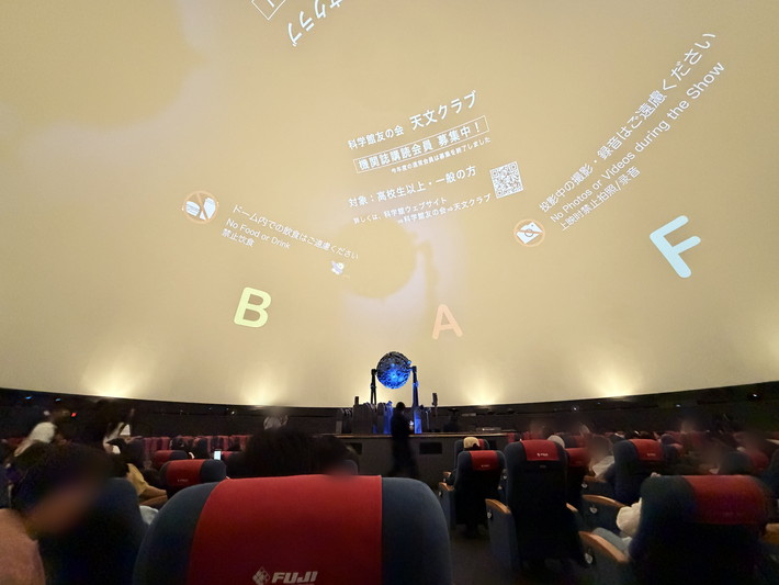 上映開始前のプラネタリウム客室内部。飲食・撮影禁止などのマナー啓発や天文クラブの広告が映し出され、静寂の中で星空に集中するための環境が整えられた、リラックスできる広々とした鑑賞空間。