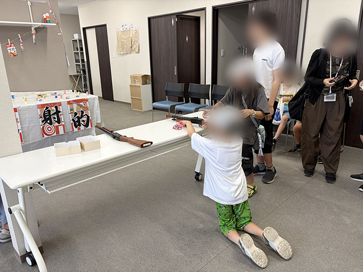 縁日気分を盛り上げる「射的」に真剣に挑戦する来場者。狙いを定めて的を射抜くドキドキ感を共有し、会場全体がどっと沸く歓声に包まれた、夏の思い出に残るにぎやかなワンシーン。