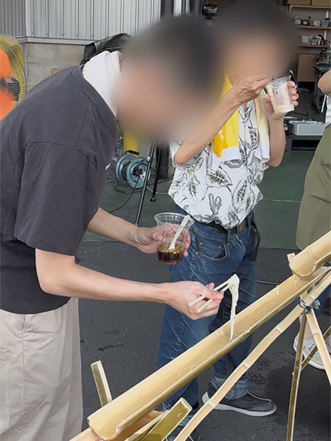 流れてくるそうめんを箸で優しくキャッチし、手元の取り皿へと移している様子。大人数が参加するイベントでの衛生面やアレルギー・感染症対策に配慮し、レーンからすくい取る箸と、口に運ぶフォークを使い分けるルールを徹底している工夫。