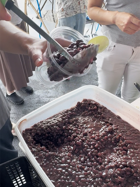デザートの「白玉あんこ(ぜんざい)」を作るために、お椀にたっぷりの甘いあずき餡をスプーンで丁寧に取り分けている様子。流しそうめんの後に、みんなで冷たくて甘い和スイーツを味わうための贅沢なデザートタイムの準備。