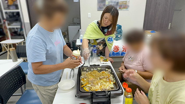 味の決め手として、具だくさんの焼きそばに塩コショウをふって味を調えている様子。訓練生とスタッフみんなで相談し合い、お好みの美味しい味付けに仕上げるこだわりの共同調理風景。