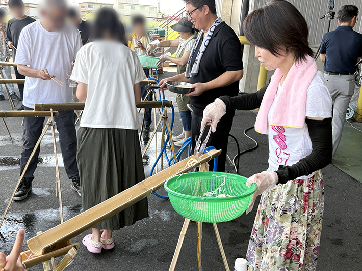 竹のレーンの上流から、訓練生たちの食べるペースに合わせて丁寧にそうめんを流していくスタッフの様子。水に乗ってさらさらと流れるそうめんを、訓練生の皆さんが笑顔で待ち構えている、レクリエーション中の温かな交流の瞬間。