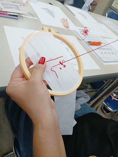 丸い木製の刺繍枠にセットした布地へ、赤い刺繍糸と針を使って真剣な表情でステッチの練習に取り組む手元写真。スタッフのアドバイスを受けながら、一つひとつ丁寧にコツを掴んで縫い進めている手芸レクの風景。