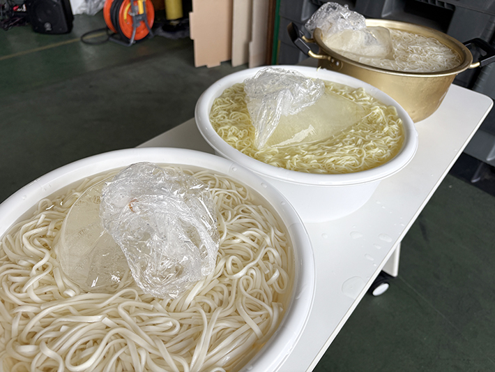 冷たい水を張った大きな容器に大きな氷の塊を浮かべ、そうめんやうどんなどの麺類をしっかりと冷やしている様子。夏の厳しい暑さの中でも、麺のコシを保ち、ひんやりと美味しく食べてもらうためのこだわりと工夫。
