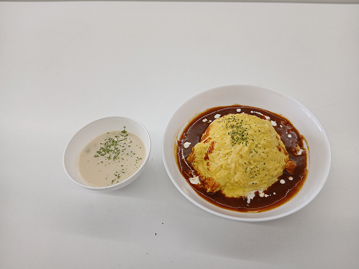 ふんわりと焼き上げた黄色い卵に濃厚なソースとパセリが綺麗にあしらわれた、特製オムライスとスープのクローズアップ。渡邉理事長のアドバイスのもと参加者が手作りで完成させた、本格的で見た目も鮮やかな自慢の逸品。