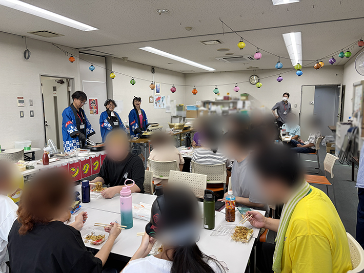 まきの木のテーブルを囲み、自分で選んだ焼きそばやたませんを「美味しい！」と笑顔で語り合いながら満喫する訓練生たちの幸せなランチタイム。