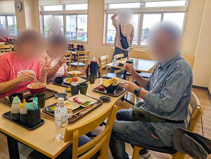 まぐろレストランの明るい室内食堂で、テーブルを囲んで仲良く昼食を味わうフレーベルの訓練生たち。テーブルの上に並べられた豪華な海鮮丼や天丼、刺身定食を前に、カメラに向かって嬉しそうにピースサインを見せる、笑顔と活気にあふれたお出かけレクのランチ風景。