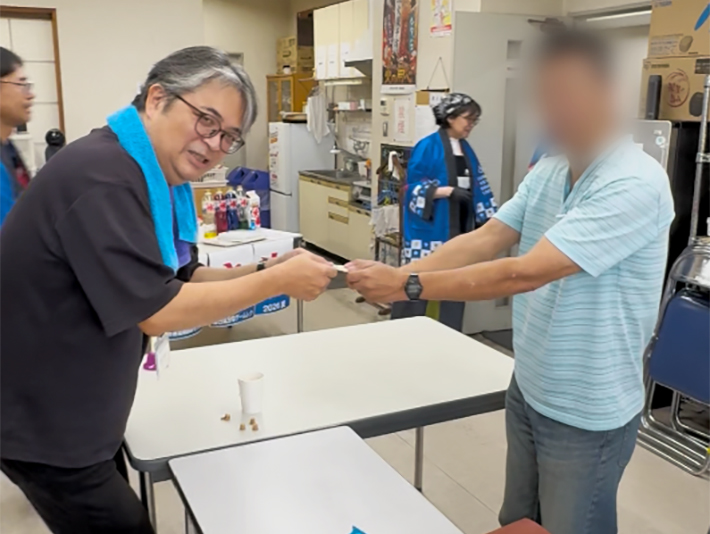 射的や千本くじで見事ターゲットを当てたり景品を引き当てたりした訓練生に、お祝いの景品を笑顔で手渡すまきの木スタッフ。ゲームの興奮と景品をもらった喜びをその場で分かち合い、信頼関係を深める温かな交流。