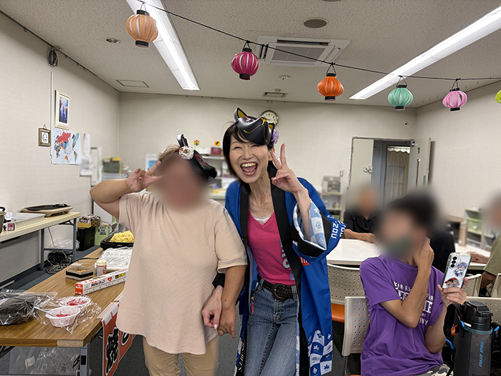 お祭り気分を一層盛り上げるために、お揃いのお面を頭に可愛らしく身につけて縁日レクを楽しむスタッフと訓練生。非日常の楽しい雰囲気に包まれてフロア全体に自然な笑顔と活気が広がる、まきの木ならではの温かな時間。