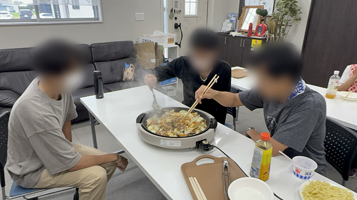 夏祭りのお楽しみであるお昼ごはん作りの様子。室内でみんなで大きなホットプレートを囲み、具材を炒めてワイワイと賑やかに焼きそば作りを楽しむフレーベルの訓練生たち。