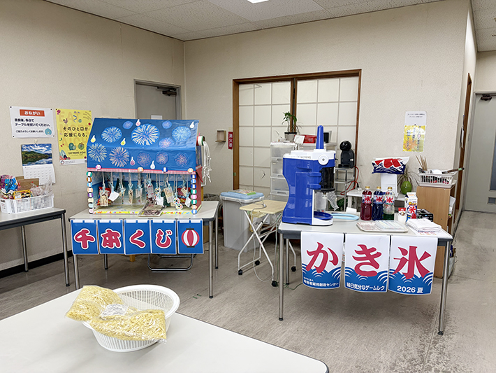 華やかな手作りのデコレーションが施された千本くじの隣に設置された、お祭りムード満点のかき氷屋台。 誰もいない静かな食堂エリアに、これから始まる賑やかな時間を予感させるように、レトロなデザインのかき氷機やシロップが整然とスタンバイしている準備風景。