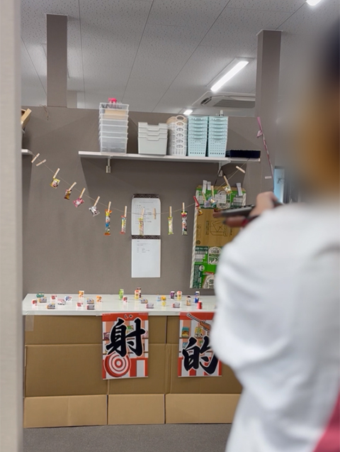 フレーベルの夏祭りゲームコーナーで、吊り下げられた的や机の上に並んだたくさんのターゲットにコルク銃の狙いを定める「射的」に挑戦する様子。得点を競い合いながら、訓練生のみんなで一緒に熱中して盛り上がる屋台ゲームの風景。