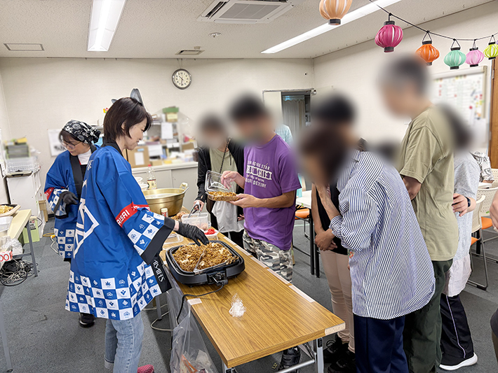 大きなホットプレートを囲み、スタッフがキャベツや麺を豪快に炒めて特製焼きそばを調理している様子。できたての香ばしい匂いが漂い、訓練生の皆さんが「美味しそう！」とお昼ごはんを心待ちにする調理風景。