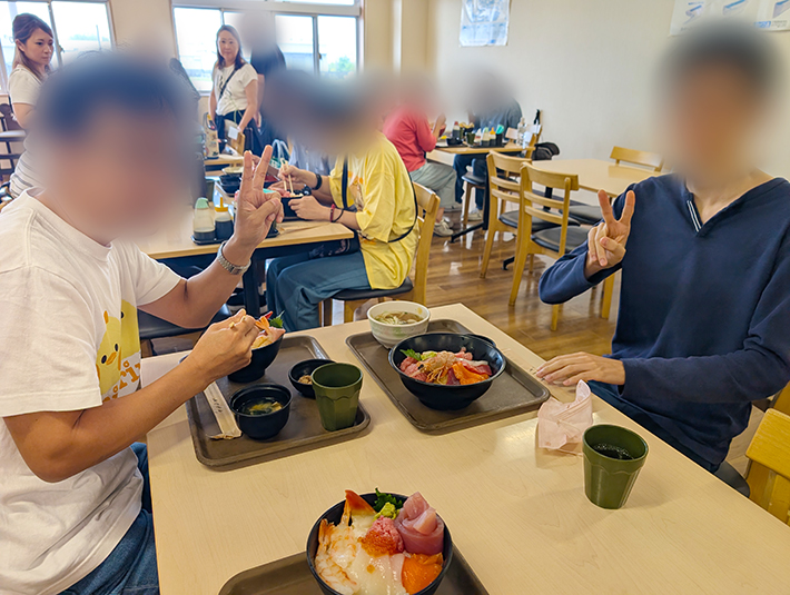 海鮮丼や海老がついた丼を囲み、両手で楽しそうにピースサインをする訓練生たちの笑顔。「どれから食べようかな」「美味しいね！」とお互いに声を掛け合いながら、みんなで一緒に食事をする楽しさと喜びを噛みしめる温かなひととき。
