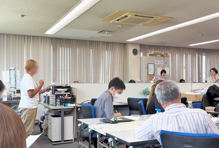 グループワークでまとまった意見や気づきを代表者が立ち上がって全体に向けて発表・共有しているシーン。他のグループの多様な視点やハッとする気づきに触れ、法人全体で支援の質向上を目指す全体共有の風景。