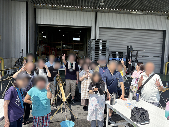イベント会場となった本部倉庫の前で、お腹いっぱいになり、水遊びも満喫して大満足の笑顔で並ぶ訓練生とスタッフ一同の集合写真。みんなの「やりたい!」が本格的な流しそうめんイベントとして形になり、最高の夏の思い出を共有した充実の記念撮影。