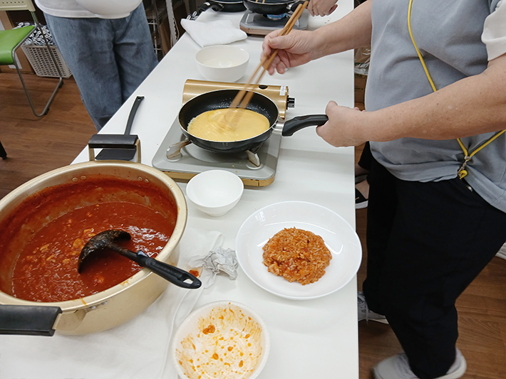 カセットコンロのフライパンで手際よく卵をかき混ぜ、自分のオムライス作りに真剣な表情で挑戦する体験風景。大鍋のソースや具材を前に、渡邉理事長からコツを教わりながら手作業で仕上げていく調理のひとコマ。