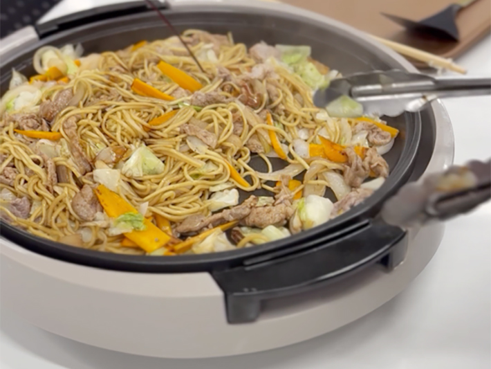 ジューシーな音を立てながら、焼きそばに特製ソースをたっぷりと加えて全体へ手際よく炒め合わせる調理工程。漂う香ばしいソースの香りに包まれ、みんなの食欲と期待が一段と膨らむランチの準備風景。
