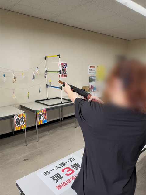 ゲームコーナーの射的で、コルク銃をしっかりと構えて吊り下げられた的を真剣に狙う訓練生。最初はなかなか当たらなくても、スタッフのレクチャーや練習を通じてコツを掴み、見事に命中させて「当たるようになってすごく楽しい！」と笑顔が弾けた瞬間。