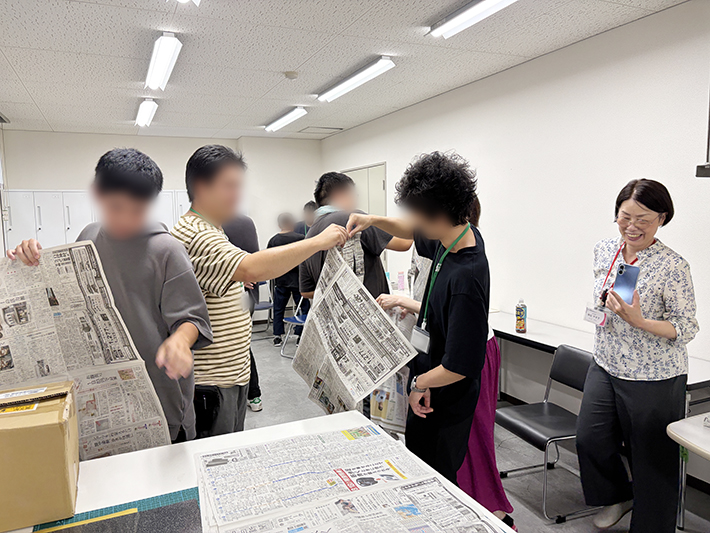 障碍者ITカレッジ愛西の就労移行支援レクリエーションで、参加者がペアになり片手で新聞紙を長く破るコミュニケーションゲームに挑戦している風景。
