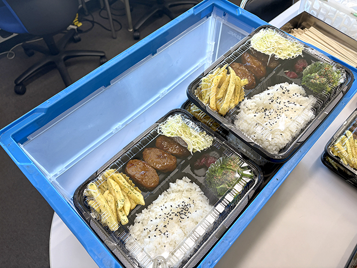 手作りお弁当を入れた青い折りたたみコンテナ(折りコン)の内部写真。コンテナの左側に大きなすき間が空いており、配送時のお弁当の傾きや崩れを防ぐための新しいスペーサー(仕切り)制作のきっかけとなった改善前の状態。