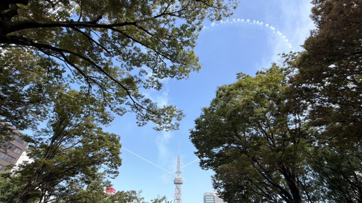 木々の向こうにテレビ塔が立ち、空にはブルーインパルスの煙の輪と交差する白い線が残る様子