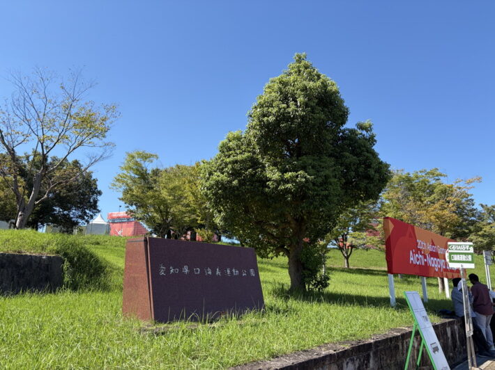 「愛知県口論義運動公園」の石碑と、芝生の斜面に立つ大会の案内看板