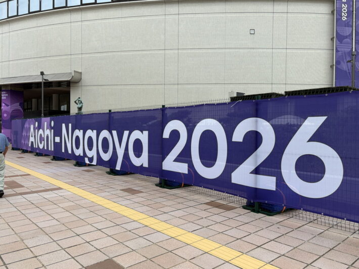 建物の壁面を覆う紫色の仮囲いに、白い文字で「Aichi-Nagoya 2026」と書かれた大会ロゴ